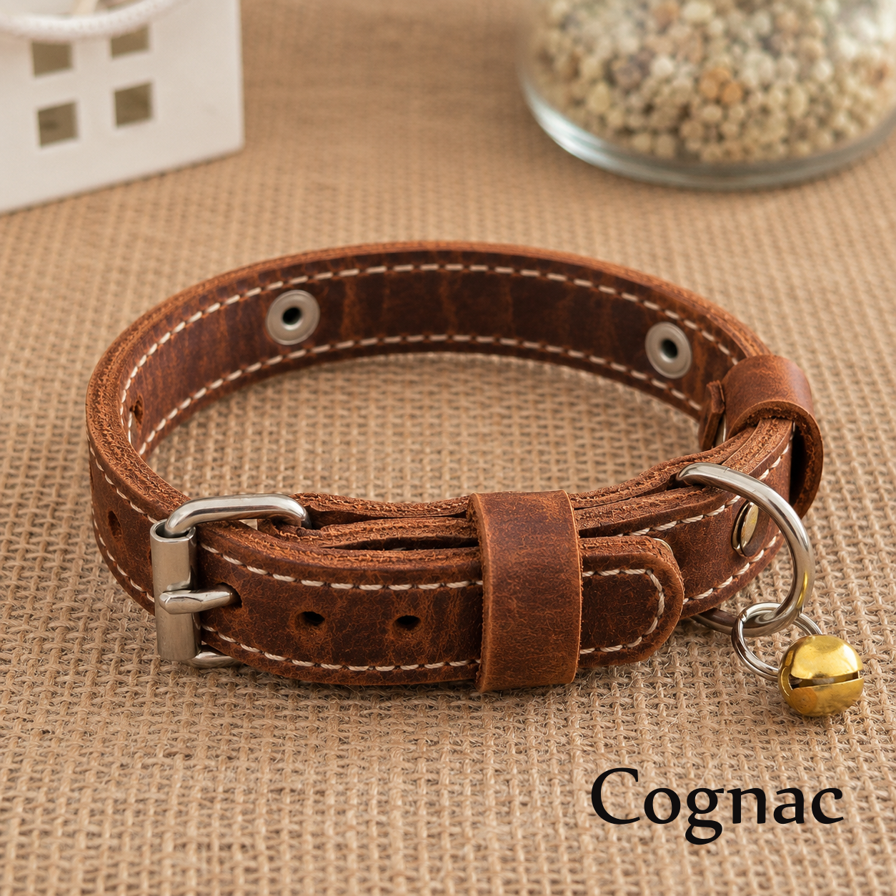 Brown leather dog collar with a gold bell on a textured surface, labeled 'Cognac'.