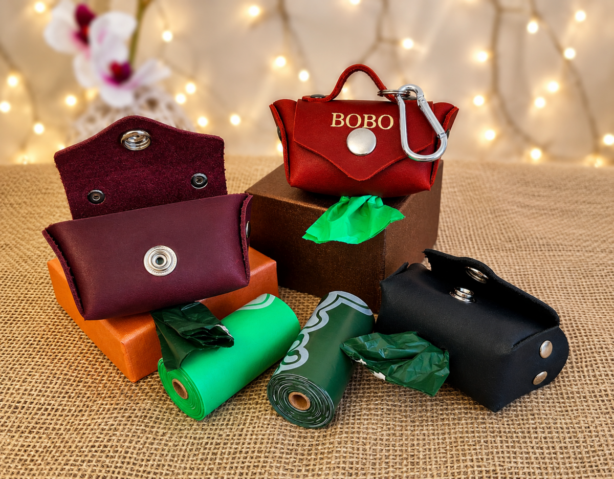 Four pet poop bags with matching zippered pouches in red, burgundy, black, and green, arranged on a burlap surface with warm string lights in the background. One red pouch has the name 'BOBO' on it.