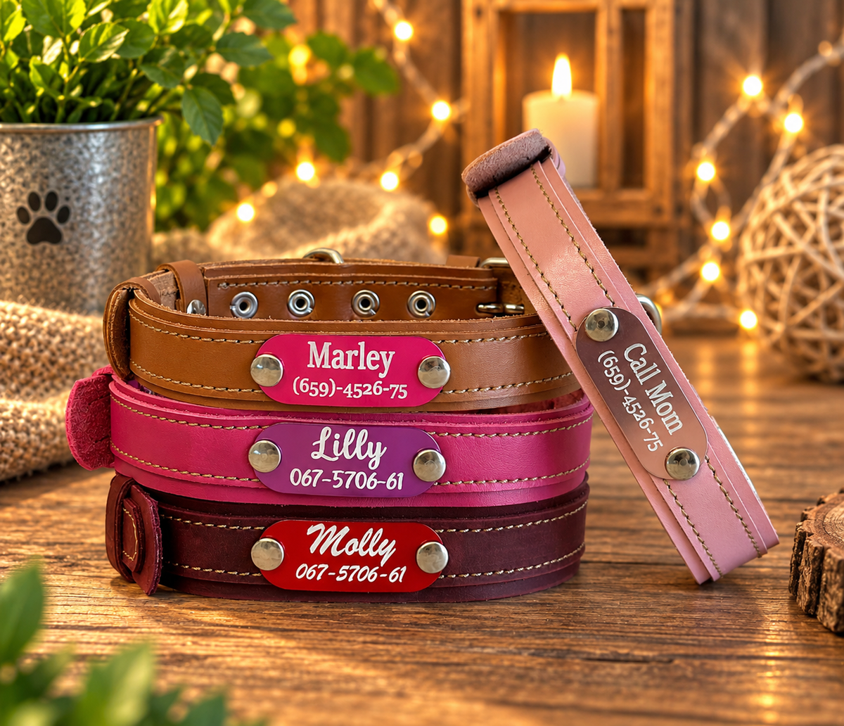 Four personalized leather dog collars stacked on a wooden table in a warm, rustic setting. The collars are brown, bright pink, dark reddish-brown, and light pink, each with a metal nameplate engraved with a pet name and phone number.