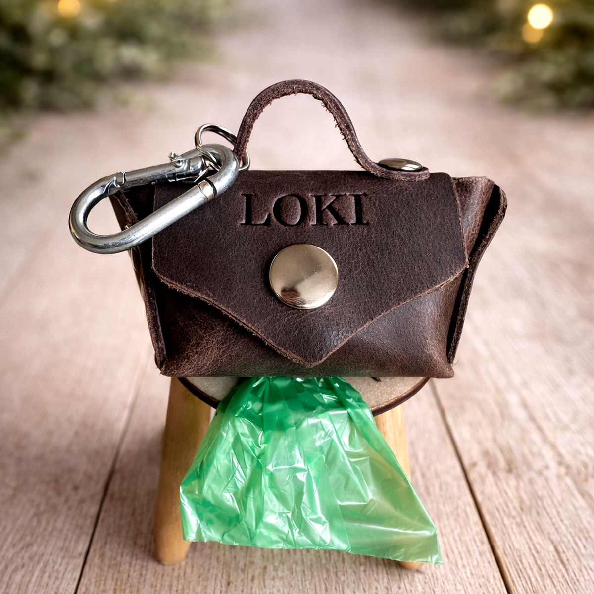 Small brown leather bag with 'LOKI' engraved on it, attached to a stand with a green plastic bag underneath.