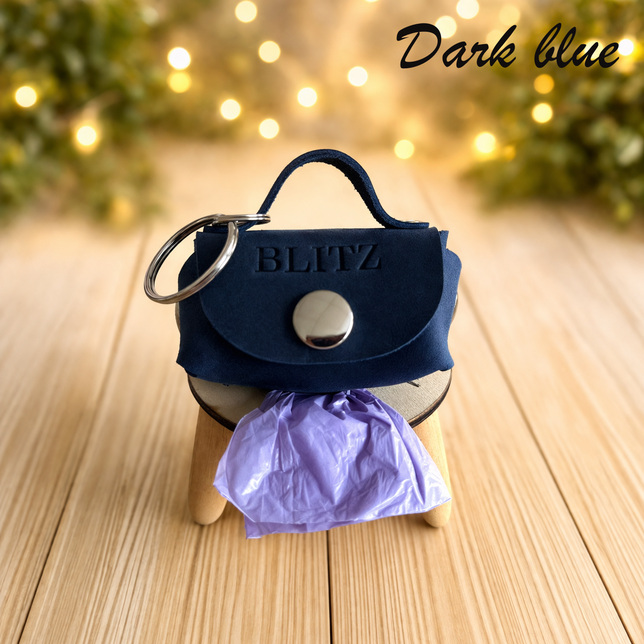 Dark blue handbag-shaped keychain with 'BLITZ' branding on a wooden surface with blurred lights in the background.