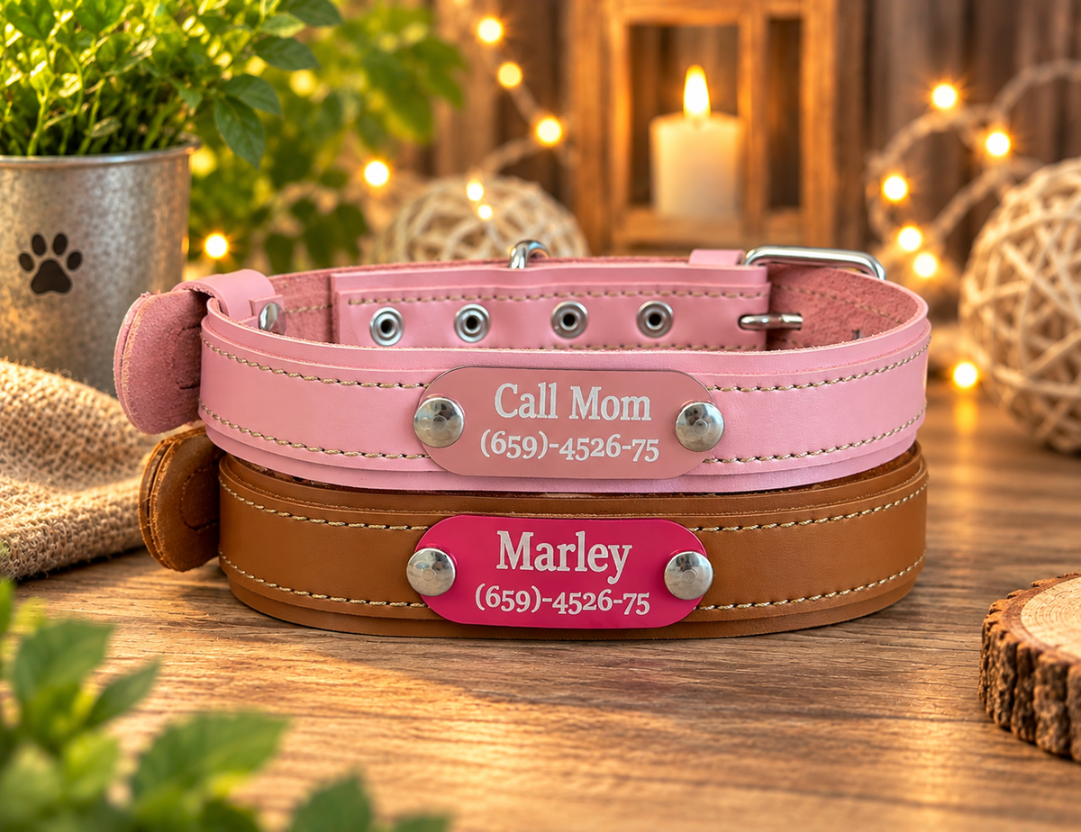 Two personalized pet collars stacked on a wooden surface: a pink collar on top with a silver plaque reading 'Call Mom (659)-4526-75' and a tan collar underneath with a matching pink plaque reading 'Marley (659)-4526-75'. The background is a warm, cozy setting with string lights, a candle, and greenery.