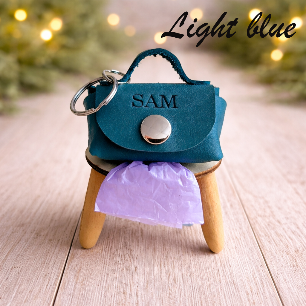 Light blue handbag-shaped keychain with 'SAM' on a small wooden stand with a blurred light background.