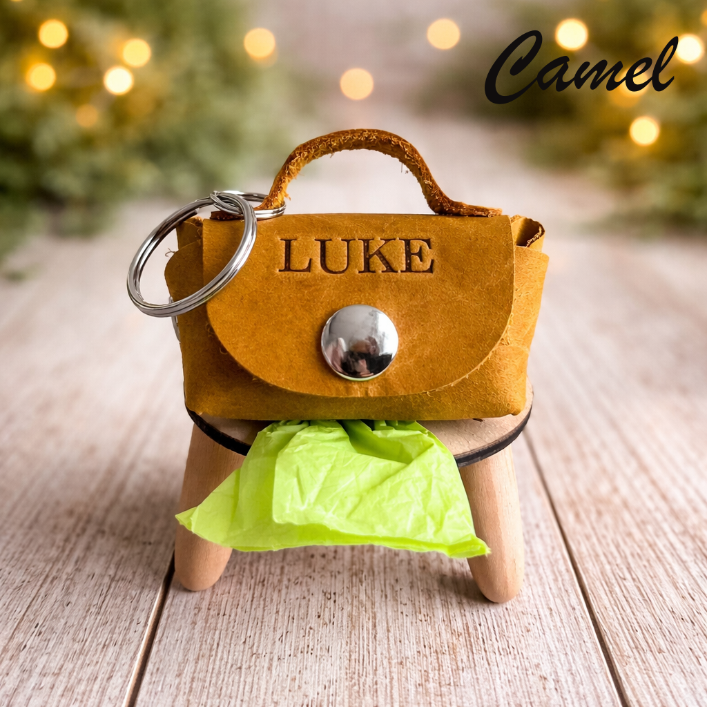 Camel-colored keychain purse with 'LUKE' engraved, on a wooden surface with blurred lights in the background.