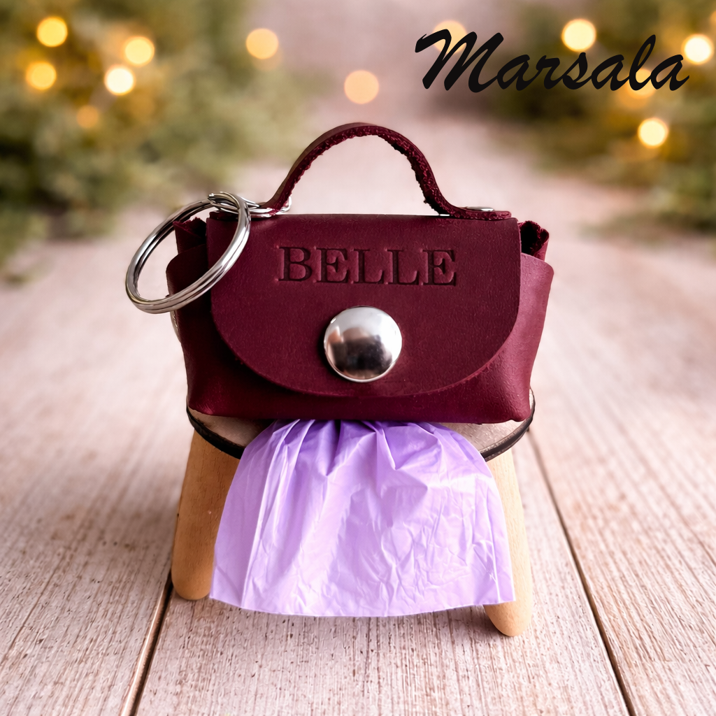 Miniature maroon handbag keychain with 'BELLE' on a stand, blurred lights in the background