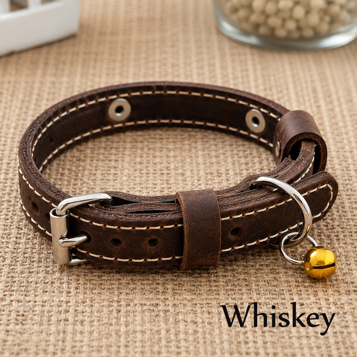 Brown leather dog collar with a gold bell on a textured surface, labeled 'Whiskey'.