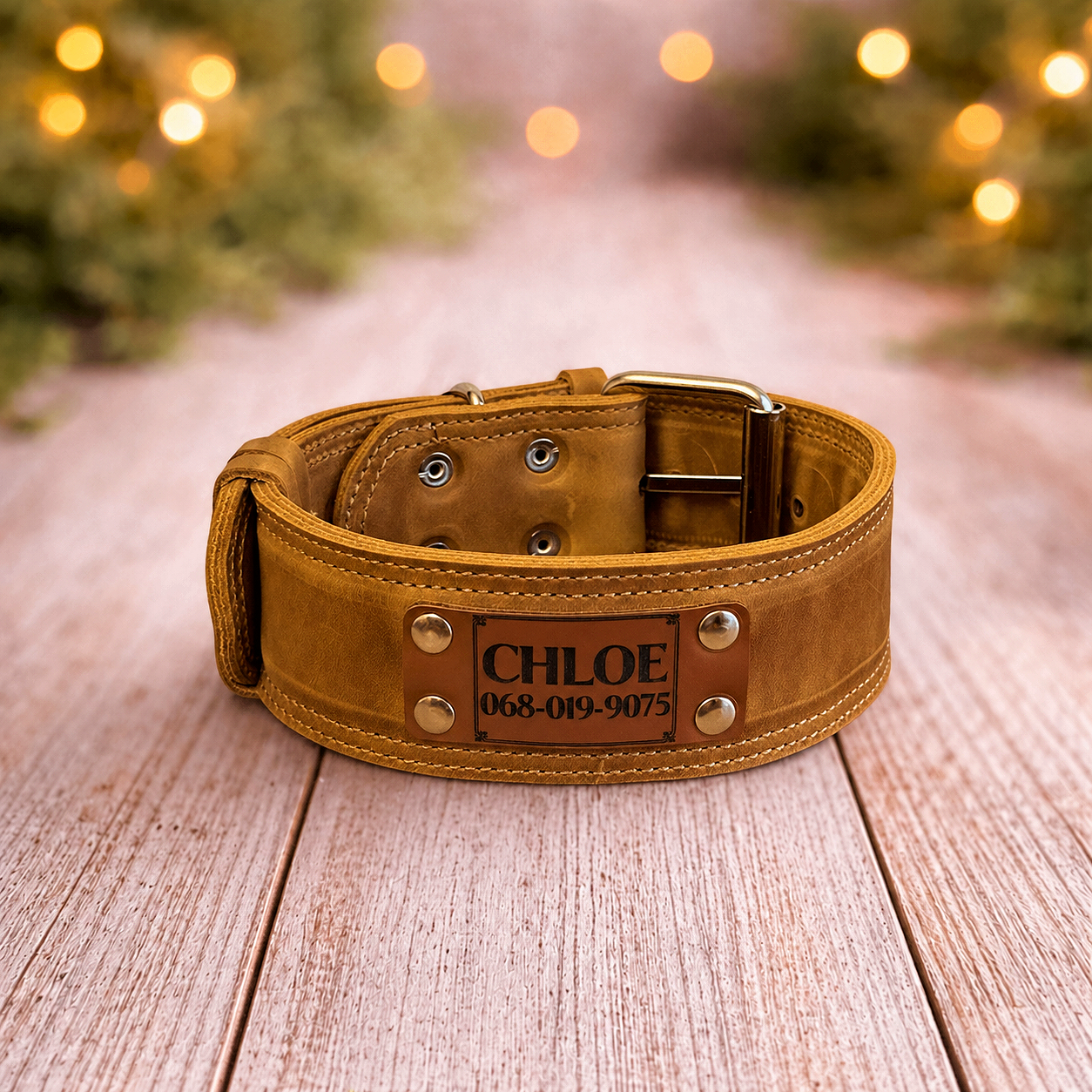Brown leather dog collar with engraved name and contact information on a wooden surface with blurred lights in the background.