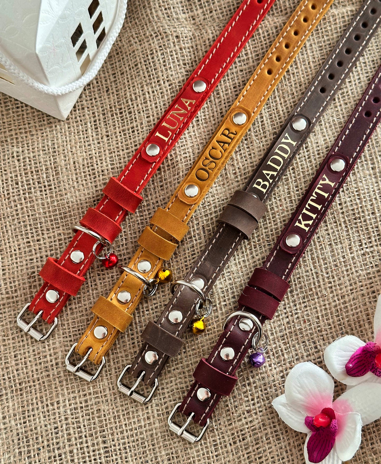 A selection of personalized leather cat collars in various colors, each featuring a nameplate. The collars are displayed on a textured background with a white bowl and purple flowers.
