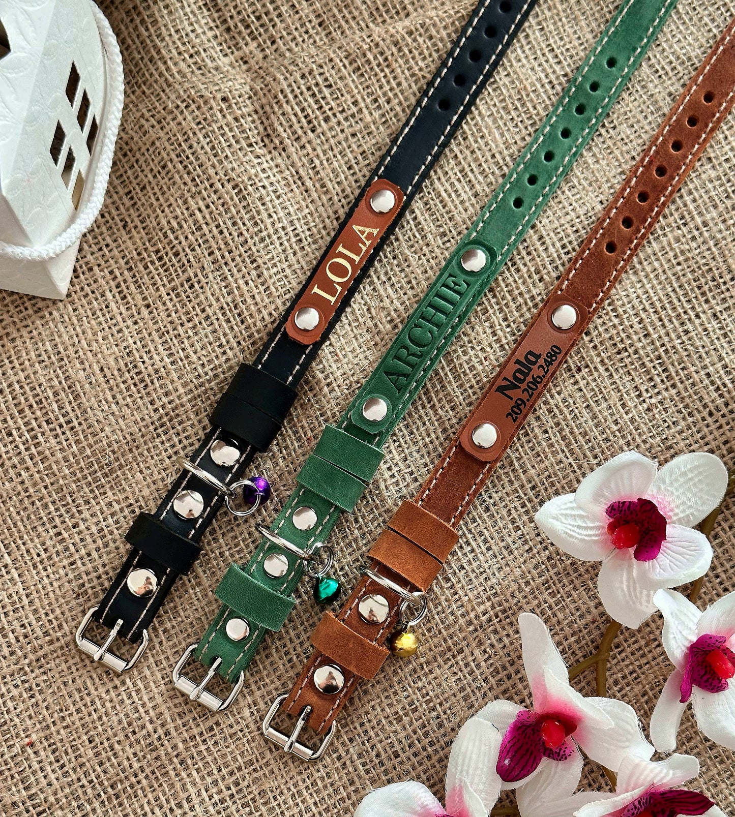 A selection of personalized leather cat collars in various colors, displayed on a textured background with flowers.