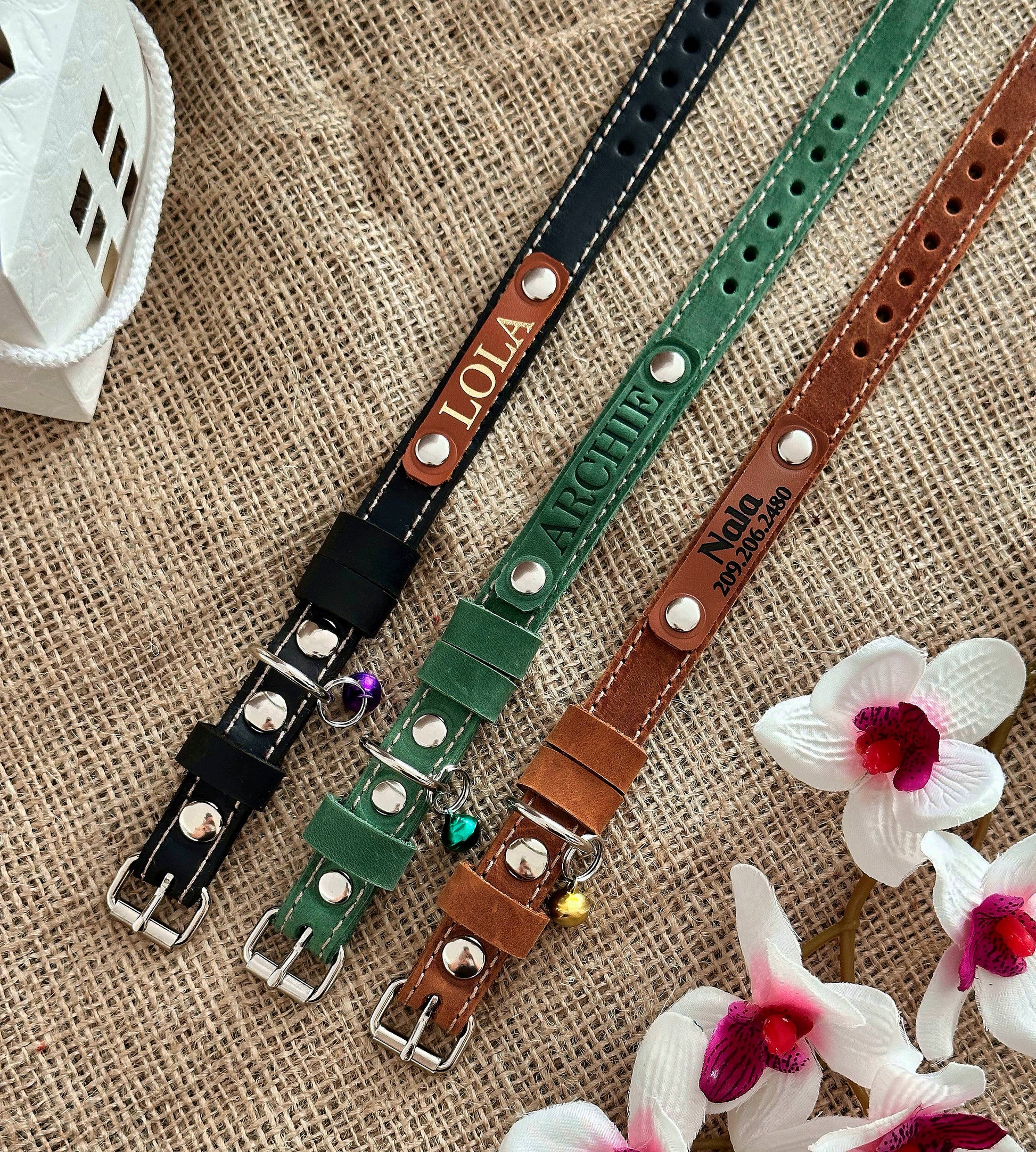 A selection of personalized leather cat collars in various colors, displayed on a textured background with flowers.