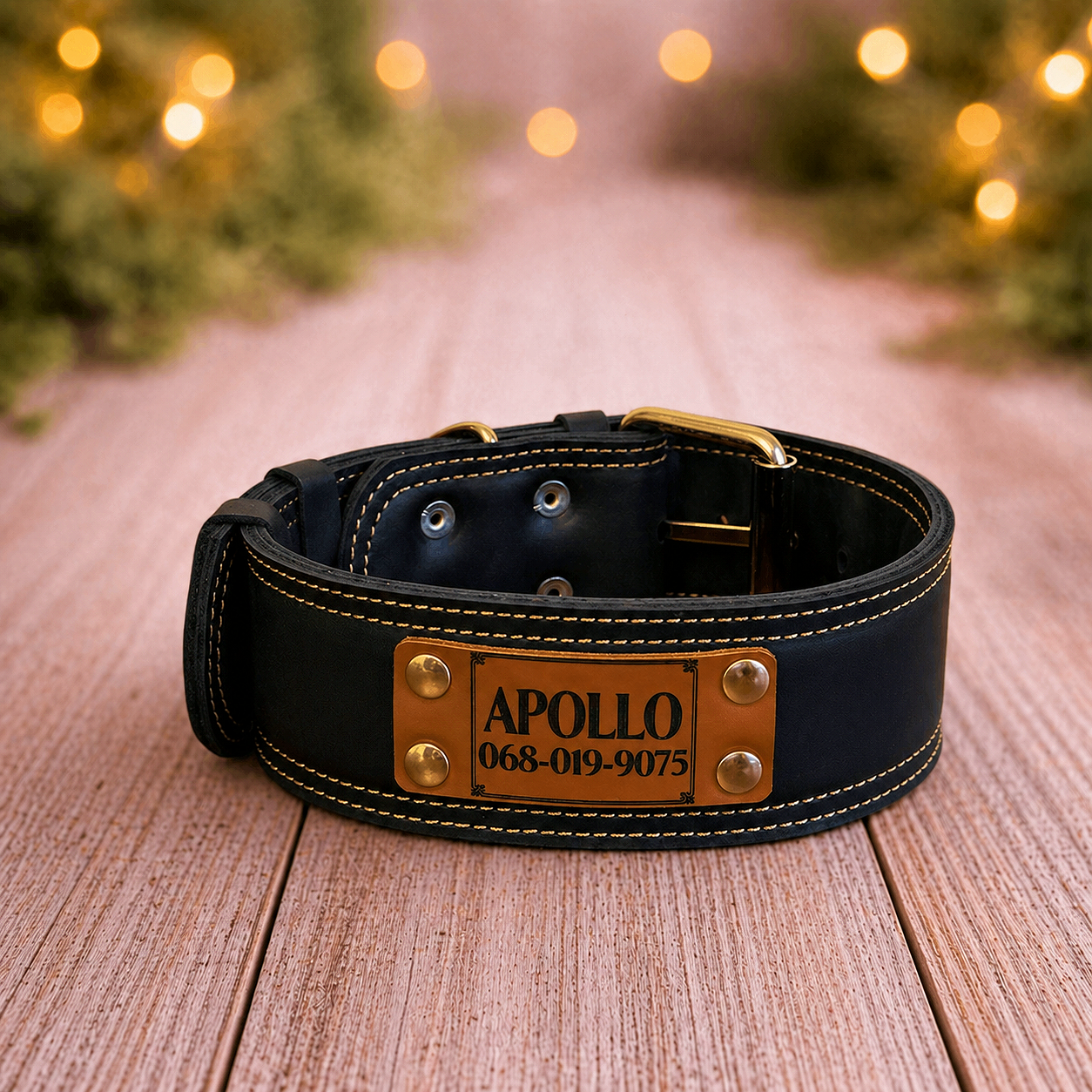 Black leather dog collar with gold hardware and engraved nameplate on a wooden surface with blurred lights in the background.