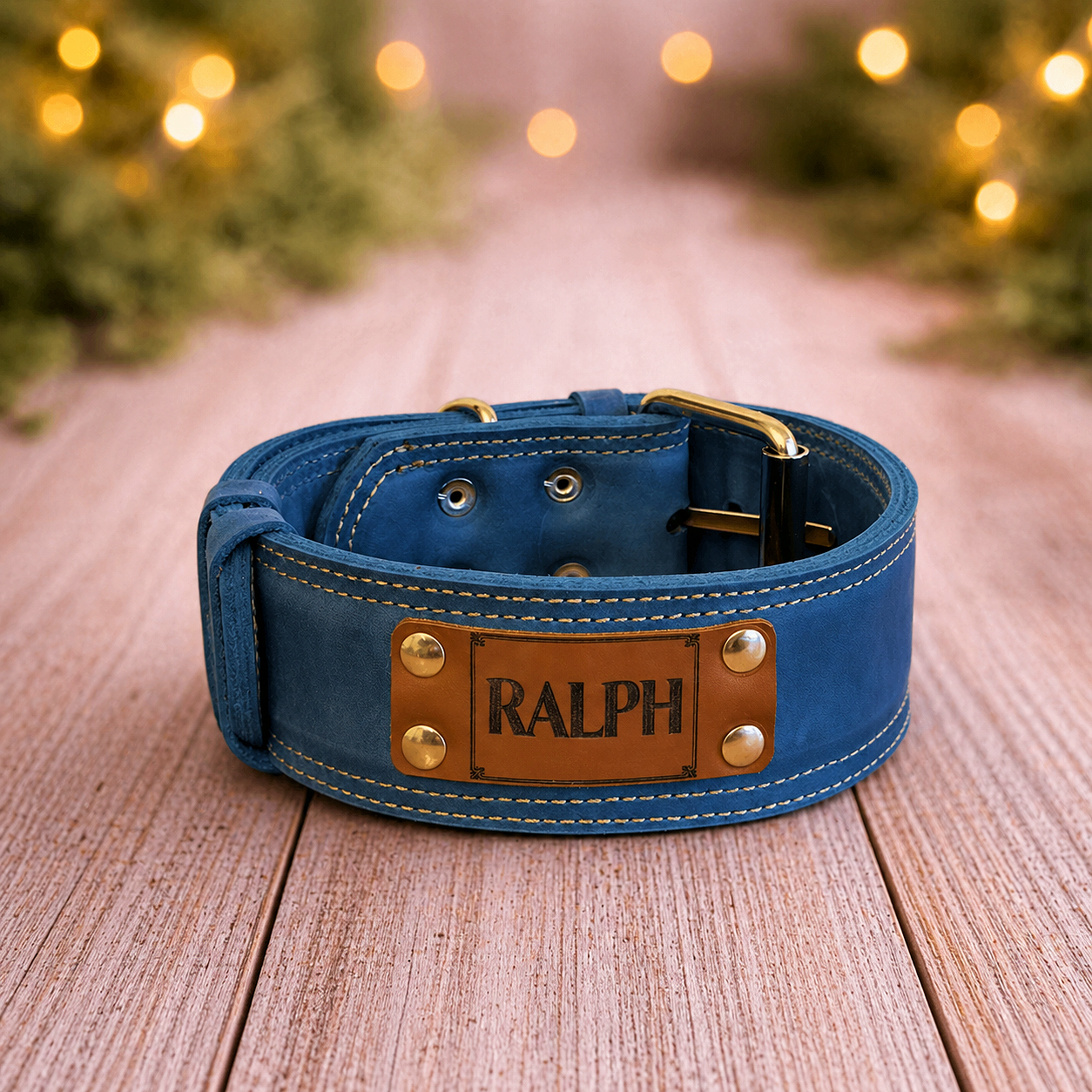 Blue dog collar with 'RALPH' label on a wooden surface with blurred Christmas lights in the background