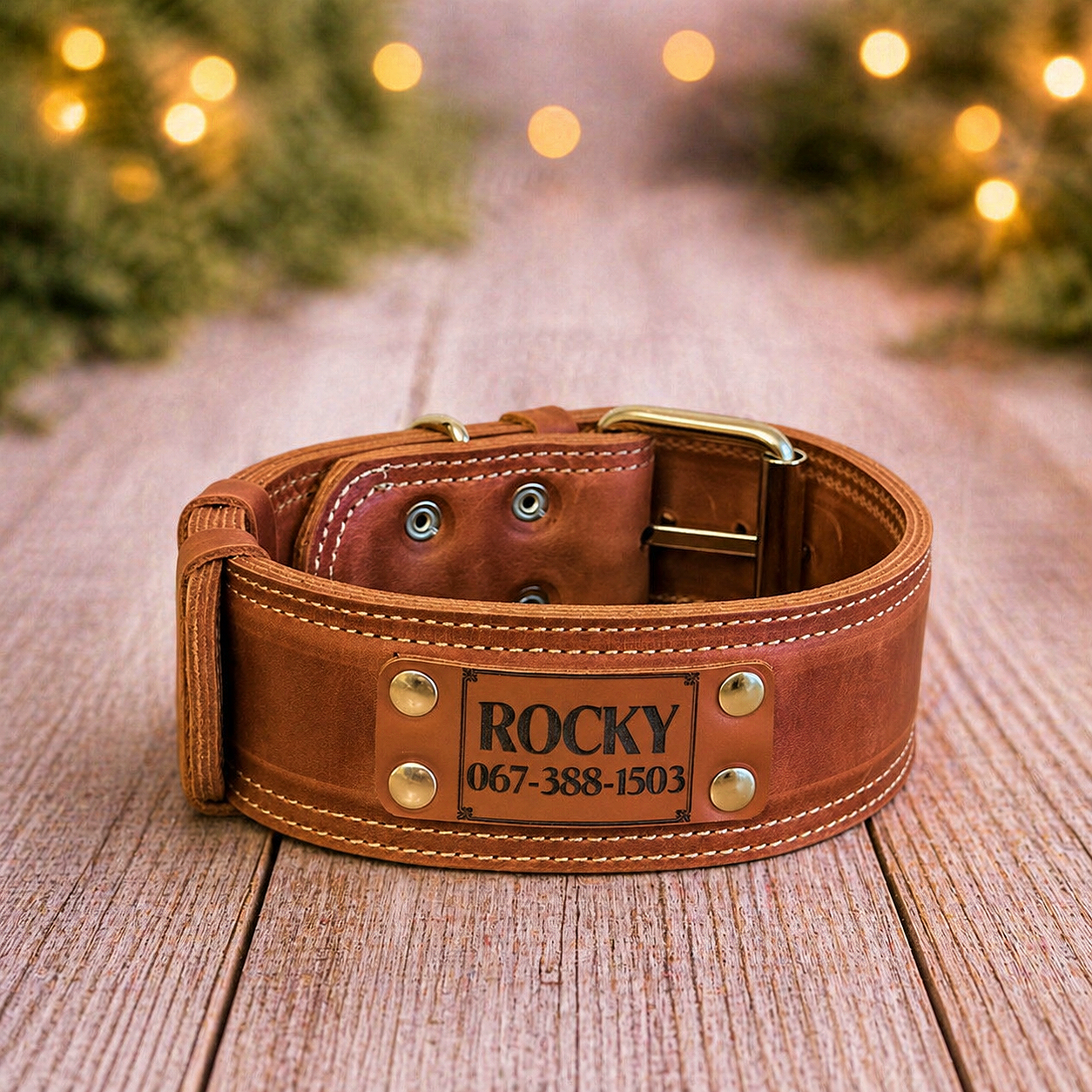 Brown leather dog collar with engraved name and phone number on a wooden surface with blurred lights in the background